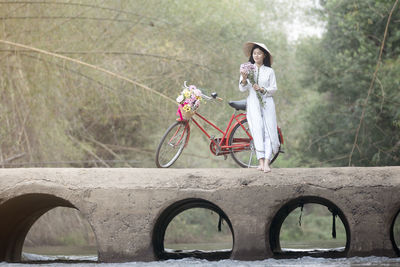 Woman leaning on bicycle while holding flowers over bridge over river