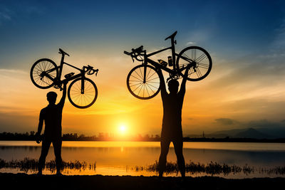 Silhouette men holding aloft bicycles standing by lake against sky during sunset