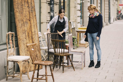 Female retailer showing antique chair to customer outside antique shop