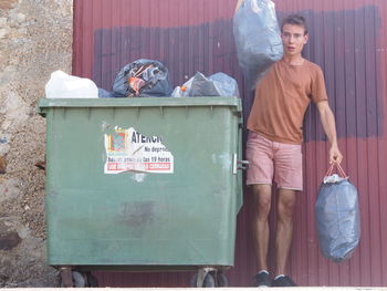Full length portrait of young man holding garbage while standing by bin