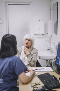 Senior female patient explaining throat problem to doctor sitting at desk in medical clinic