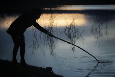 Silhouette woman holding stick in lake during sunset