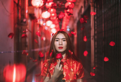 Portrait of young woman holding red rose while standing in alley at night