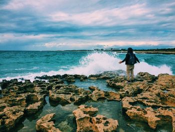 Rear view of man standing by waves splashing on rocks against cloudy sky