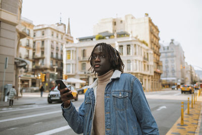 Young man looking away while holding smart phone standing in city