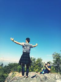 Man photographing friend standing on rock against blue sky