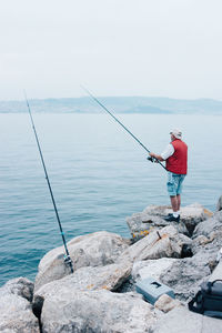 Side view of active man in red waistcoat with fishing rod standing on stony shoe and fishing in bright day