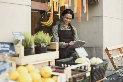 Smiling female owner using digital tablet while sitting at store entrance