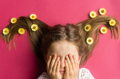 Directly above shot of girl with flowers on hair hiding face with hands over pink background