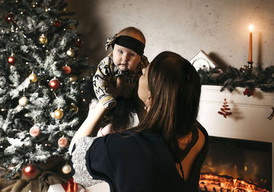 Woman holding baby girl by christmas tree at home