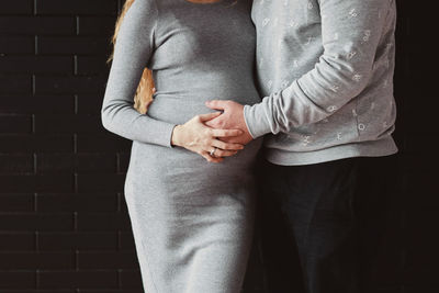 Midsection of man and pregnant woman with hands on stomach standing against wall