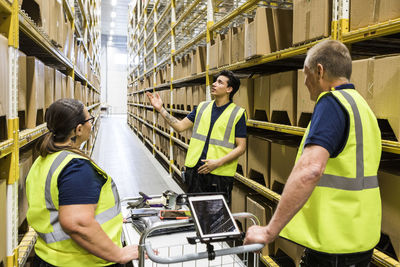 Young worker discussing with coworkers with cart while standing on aisle amidst racks at distribution warehouse