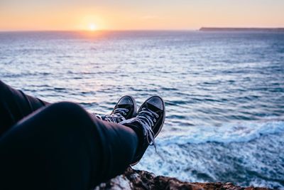 Low section of man sitting on cliff by sea during sunset