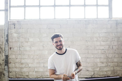 Portrait of confident smiling young businessman standing with smart phone against brick wall at workplace