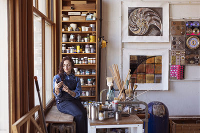 Female artist using mobile phone while sitting by window in workshop