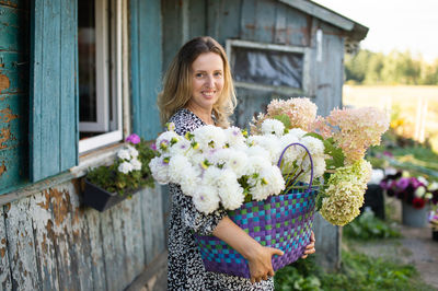 A girl in a dress with a basket of dahlia flowers stands near a house in the village and smiles