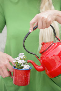 A blonde with long hair in a bright green dress pours water from a red teapot into a red iron mug 