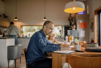 Side view of male freelancer writing in book while sitting near dining table at home
