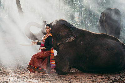 Portrait of smiling woman sitting with elephant in forest