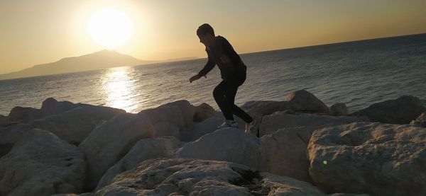 Man standing on rock by sea against sky during sunset