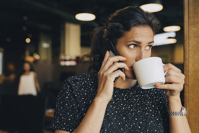 Mid adult businesswoman drinking coffee while using smart phone in office