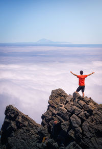 Back view of anonymous male traveler standing with outstretched arms on rocky hill in highlands and enjoying amazing view of clouds
