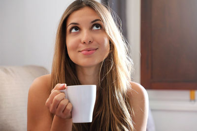 Thinking concerned young woman looking up with cup of coffee on couch at home