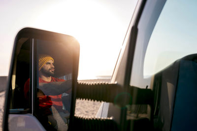 Reflection in side view mirror of male explorer sitting on driver seat in camper parked on beach and admiring seascape during summer trip