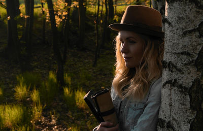 Thoughtful young woman with book standing by trees in forest