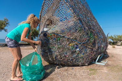 Mature blonde woman putting a plastic bottle into a trash can full of recyclable material.