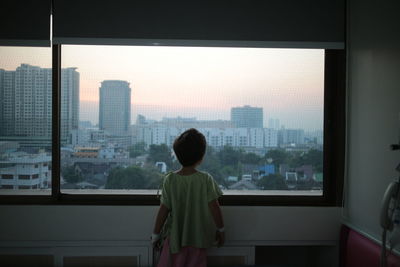 Rear view of boy looking through window at cityscape