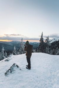 Man photographing while standing on snow covered landscape during sunset