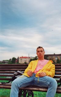 Portrait of young man sitting on bench against cloudy sky