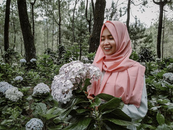 Smiling woman wearing hijab standing by flowering plants in forest
