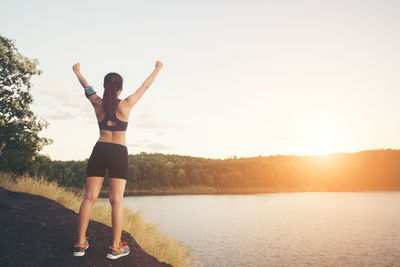 Rear view of successful female athlete standing with arms outstretched on rock at lakeshore during sunset