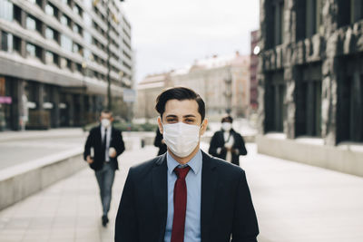 Male entrepreneur with colleagues in background walking on footpath by office building during covid-19