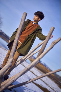 Young man looking away while standing by wooden posts against sky