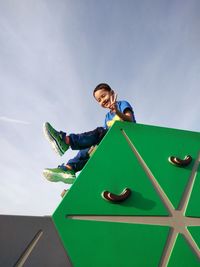 Low angle portrait of boy gesturing while sitting on roof against sky