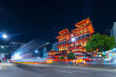 Illuminated city street by buildings against sky at night