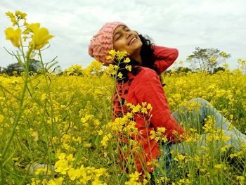 Smiling woman sitting amidst flowers on field against sky