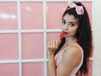 Portrait of young woman holding lollipop against pink wall