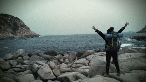 Rear view of man standing on rock at sea shore against sky