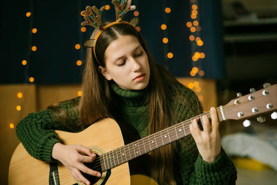 A beautiful teenage girl in christmas antlers and a green sweater plays the guitar