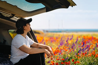 Side view of young man standing in suv car  by flowering wild plants in summer