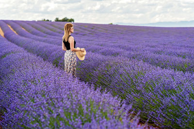 Woman in long dress and sunglasses standing in purple blooming lavender flowers field looking away.