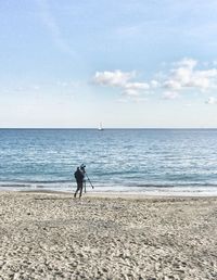 Rear view of man with tripod and camera walking at beach against sky