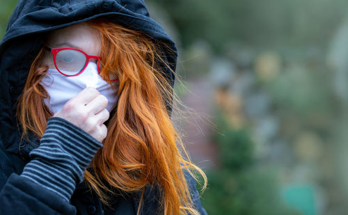 A hooded woman with red hair stands lonely and sad in a park. she wears a breathing mask.