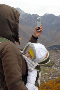 Side view of woman using phone while standing with baby against mountains