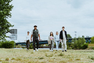 Full length portrait of young friends walking towards river against sky