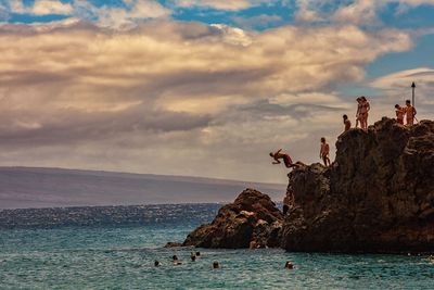 People on rock formation by sea against cloudy sky